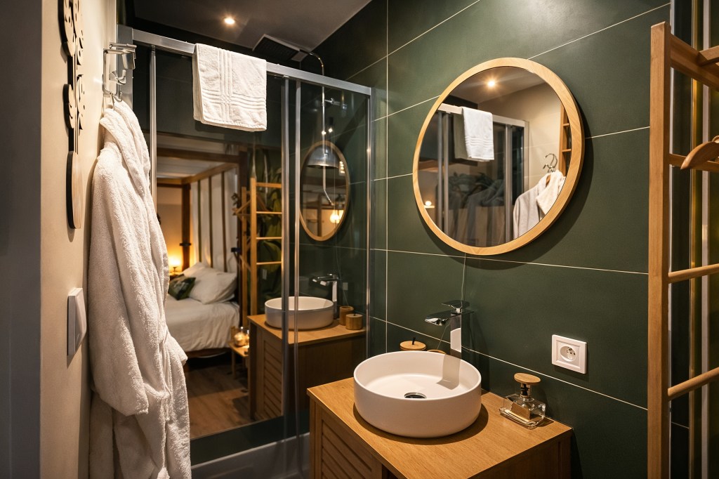 Salle de bain moderne avec vasque en céramique, meuble en bois, miroir rond et peignoirs dans un hébergement avec jacuzzi à Angoulême